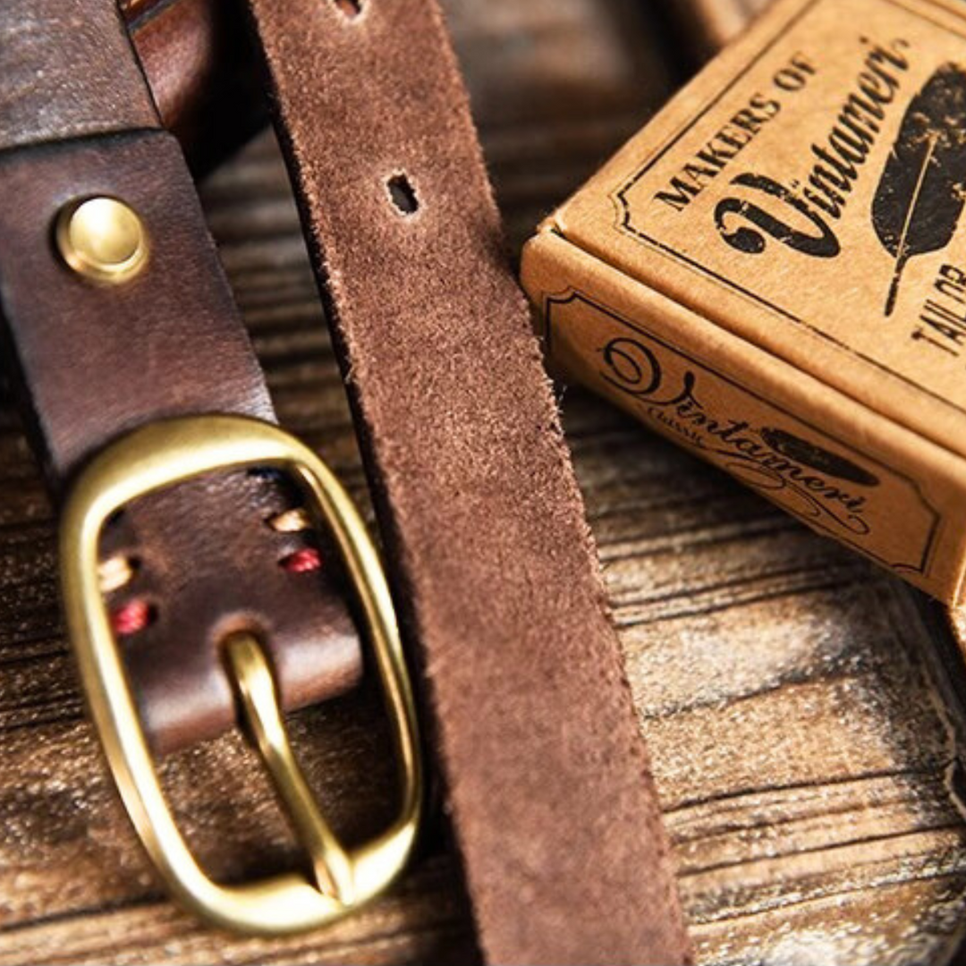 Close-up of a brown leather belt with a brass buckle, perfect for country fashion, lying next to a vintage-style box labeled Makers of Vintagework. The belt features visible stitching and punched holes.