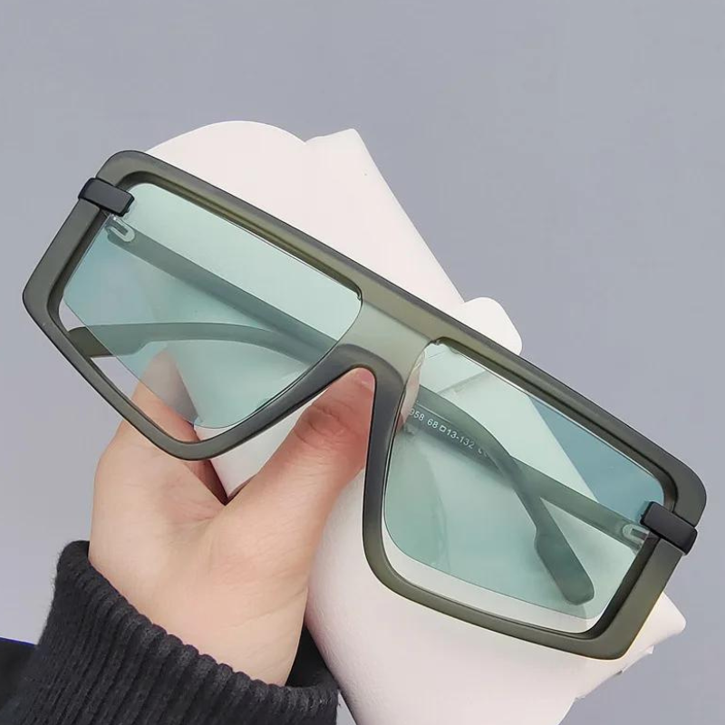 A hand holds a pair of oversized, rectangular green sunglasses with light blue tinted UV400 lenses and thick polycarbonate frames against a plain gray background, ensuring both style and sun protection.