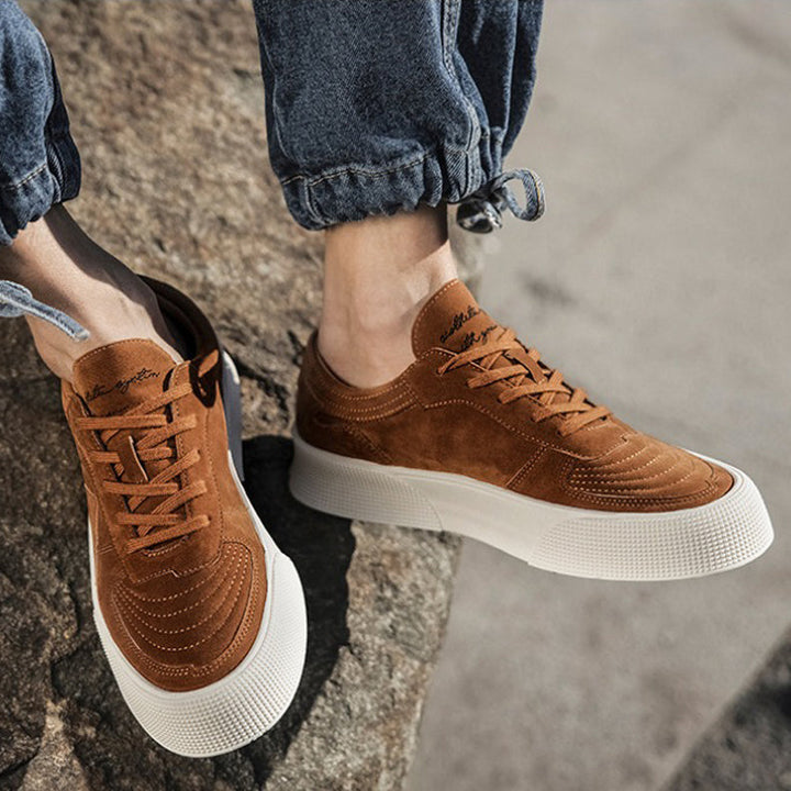 A person wearing light brown suede lace-up sneakers with comfortable cushioning and white soles, paired with blue cuffed jeans, stands outdoors on a stone surface in bright sunlight.
