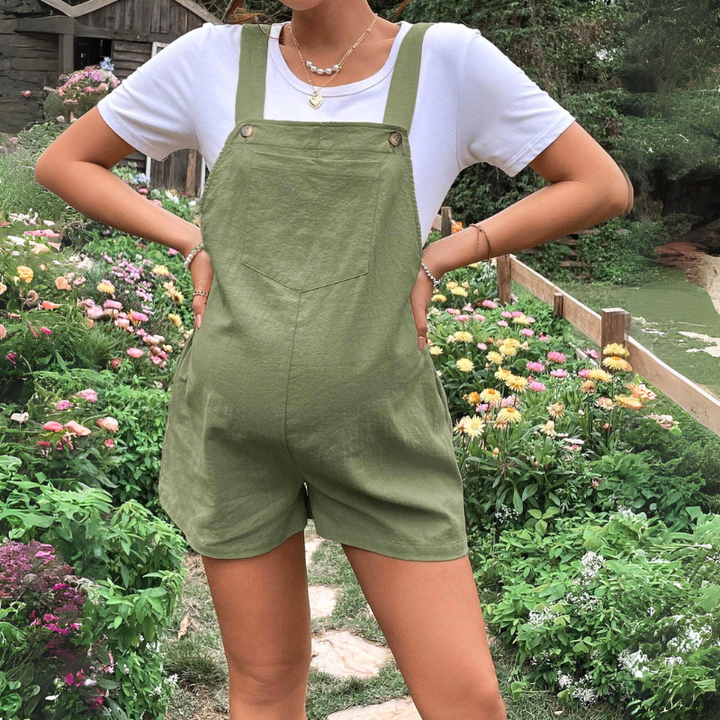 A woman wearing green pregnancy overalls and a white T-shirt stands in a garden filled with colorful flowers and greenery, her hands on her hips. Her face is not visible.