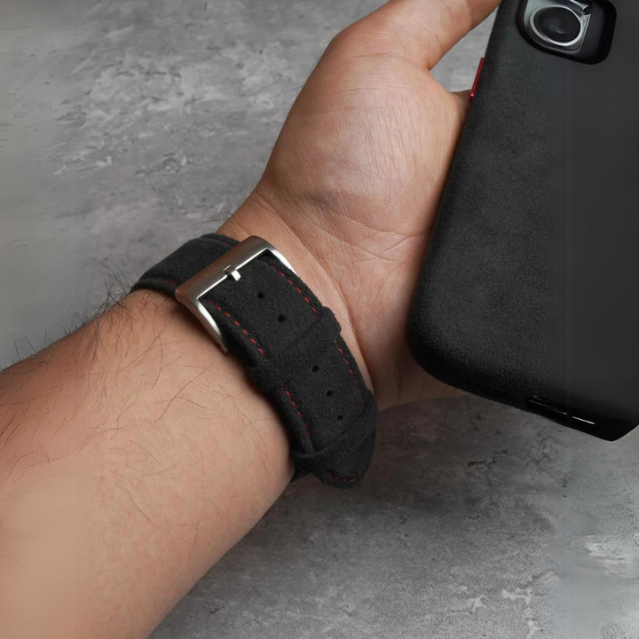A person wearing a black suede watch strap with red stitching and a stainless steel buckle is holding a black smartphone in their hand over a gray, textured surface.