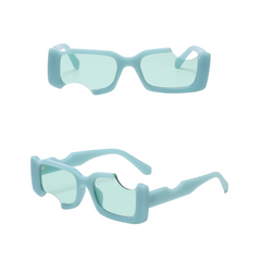 Two views of avant-garde sunglasses featuring light blue, thick frames and rectangular, green-tinted lenses with a distinctive semicircular cutout on the left lens. Shown from the front and at an angle, these irregular sunglasses offer UV400 protection.