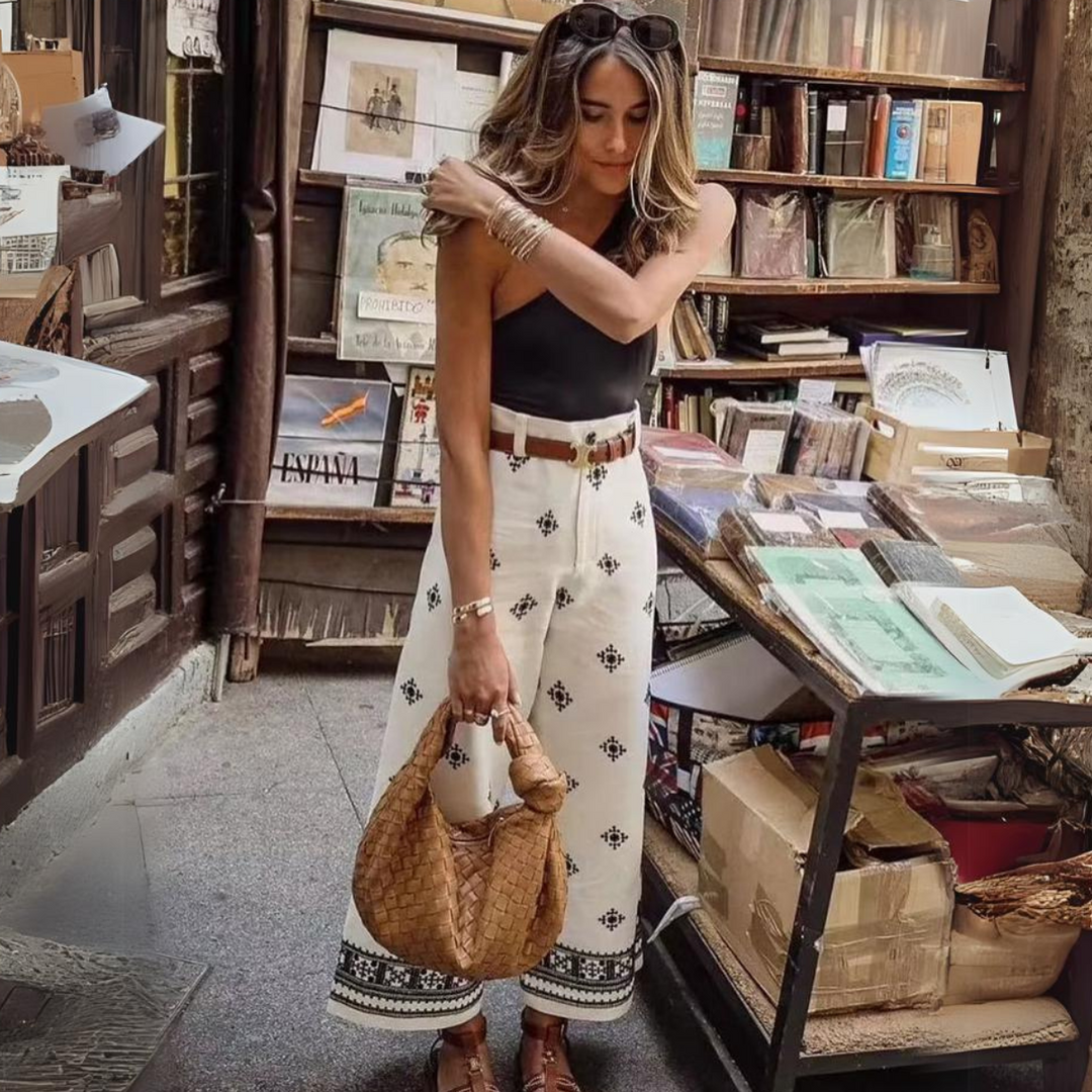 A woman with long hair, wearing sunglasses, a black top, wide-leg trousers, and brown sandals, stands near a bookstall outdoors, holding a woven bag and looking down.