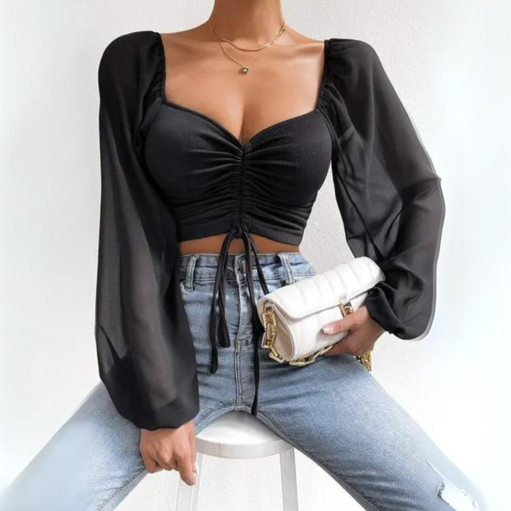 A woman wears a black, long-sleeve mesh crop top with a ruched front and flirty neckline, paired with light blue jeans. She sits on a white stool holding a small white handbag against a plain white background.