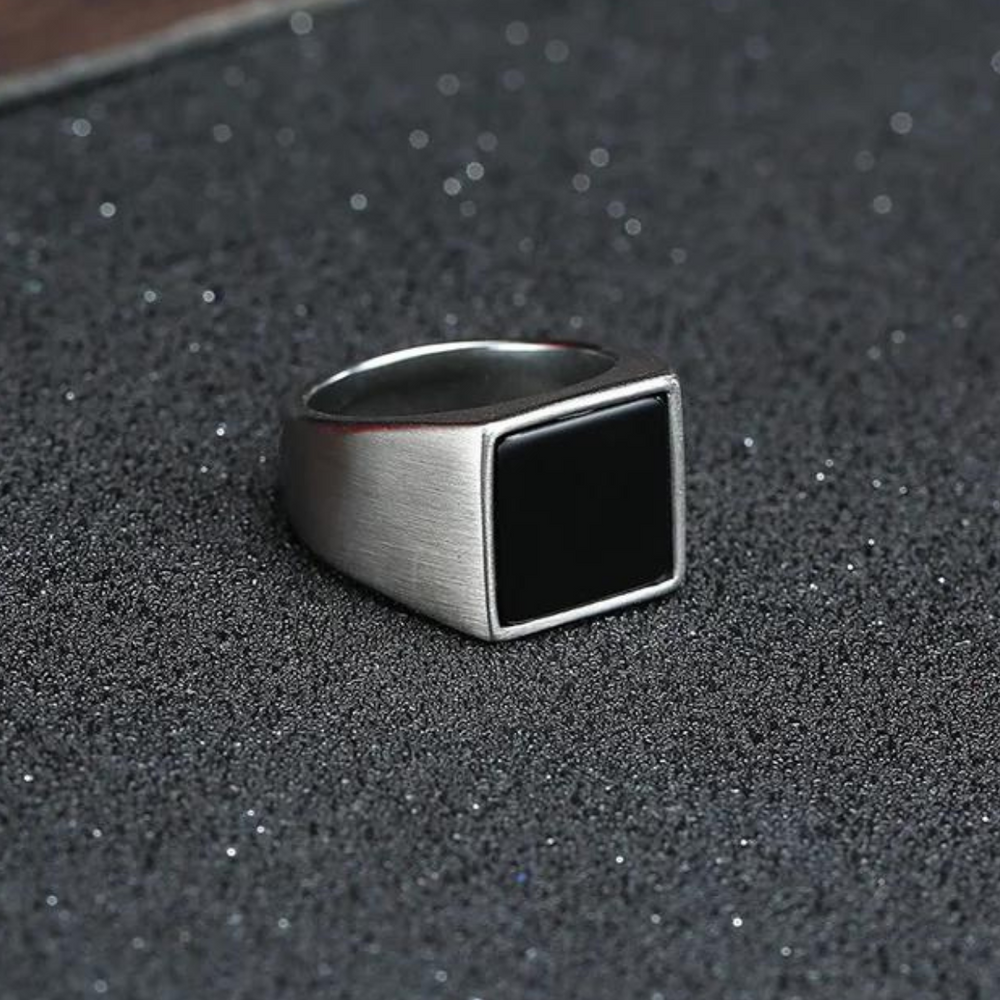 A silver ring with a large, flat, black rectangular face and a polished finish rests on a textured, dark surface sprinkled with fine glitter.