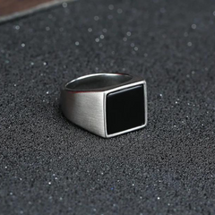 A silver ring with a large, flat, black rectangular face and a polished finish rests on a textured, dark surface sprinkled with fine glitter.