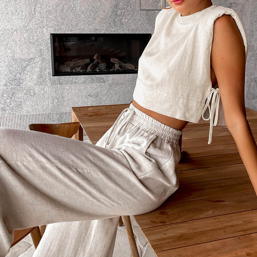 A person wearing a light beige Belgian Linen Blend cropped top with side ties and matching wide-leg pants sits on a wooden table in a modern indoor setting with a stone wall and built-in fireplace in the background.