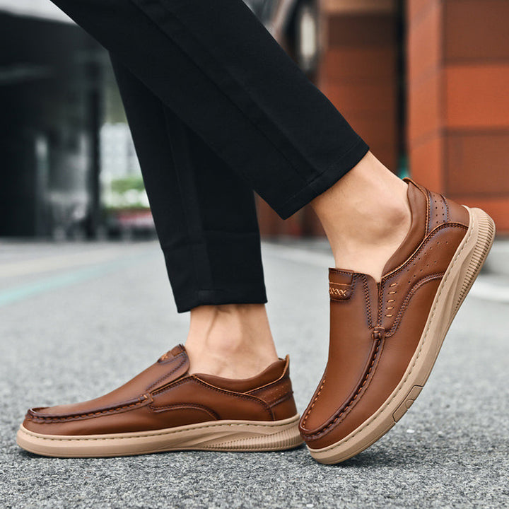A person wearing slip-on men's cowhide leather shoes with soft soles stands on a city sidewalk. The brown loafers complement black pants, while buildings create a blurred urban backdrop.