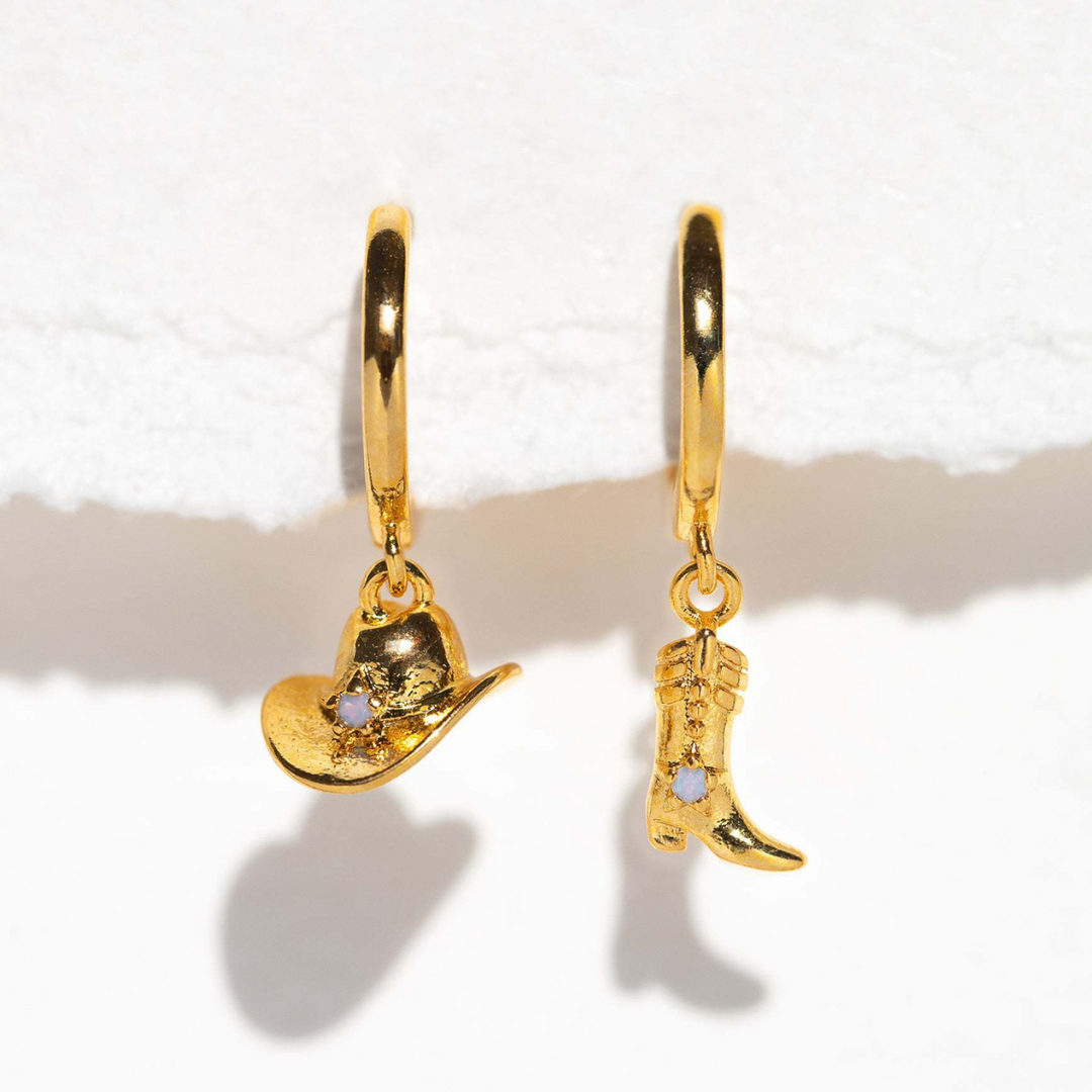 A pair of statement earrings: gold hoops, one with a cowboy hat charm and the other with a cowboy boot charm, each adorned with a small clear gemstone, displayed on a white surface.