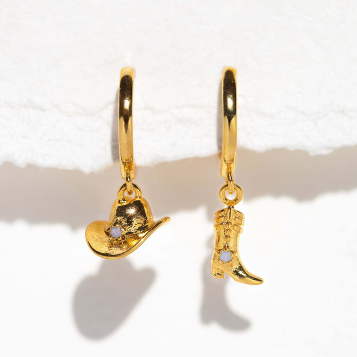 A pair of statement earrings: gold hoops, one with a cowboy hat charm and the other with a cowboy boot charm, each adorned with a small clear gemstone, displayed on a white surface.