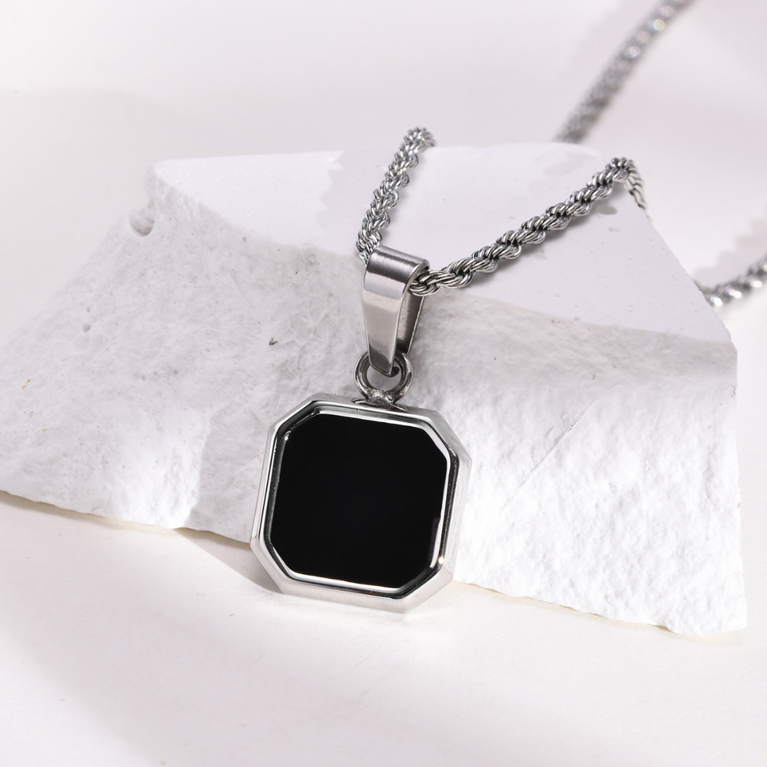 A silver necklace with a square Onyx Stone Amulet pendant rests on a white, textured stone surface. The chain is rope-style, and the collectable jewelry piece features a shiny metal frame with beveled edges.