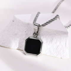A silver necklace with a square Onyx Stone Amulet pendant rests on a white, textured stone surface. The chain is rope-style, and the collectable jewelry piece features a shiny metal frame with beveled edges.