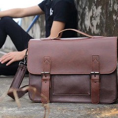 A brown premium leather satchel with buckle straps sits on the ground in focus, while a person in black clothing sits blurred in the background against a weathered wall.