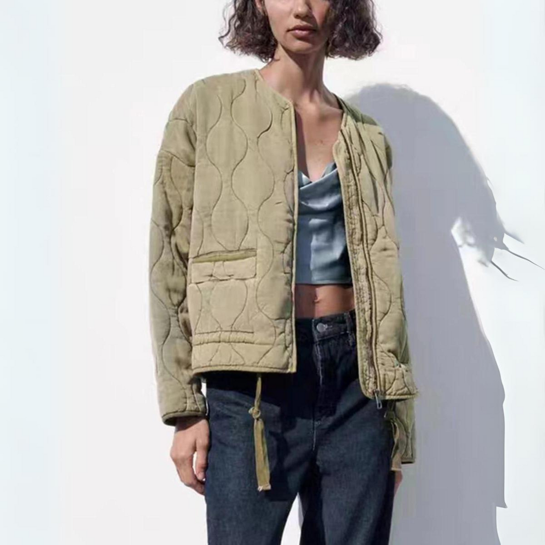 A woman wearing a light khaki quilted bomber jacket with padded insulation over a light blue satin crop top and dark jeans stands against a white background, partially shadowed.