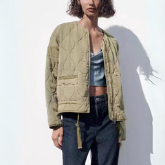 A woman wearing a light khaki quilted bomber jacket with padded insulation over a light blue satin crop top and dark jeans stands against a white background, partially shadowed.