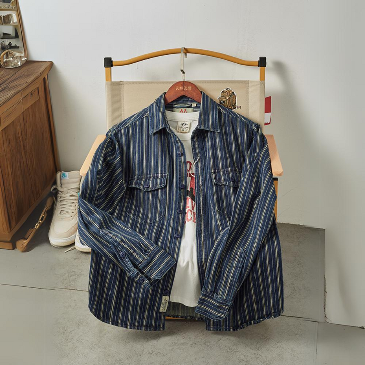 A blue and yellow vertical striped denim jacket is draped over a chair, with a white graphic T-shirt underneath. A white sneaker rests on the floor beside the wooden cabinet next to the chair.