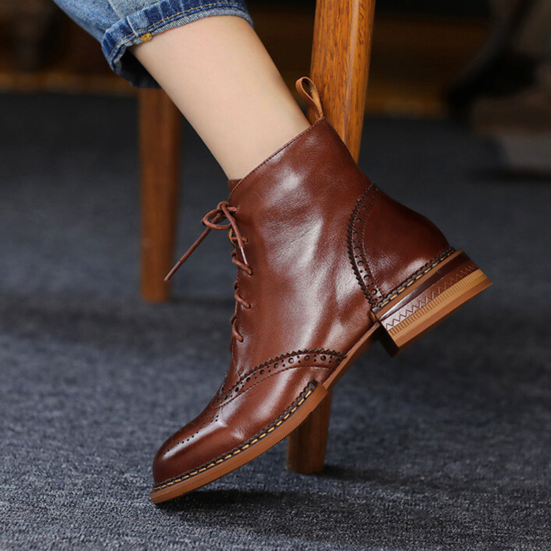 A person wearing blue jeans and genuine leather brown lace-up ankle boots rests their foot on the leg of a wooden chair, with a dark carpeted floor beneath.
