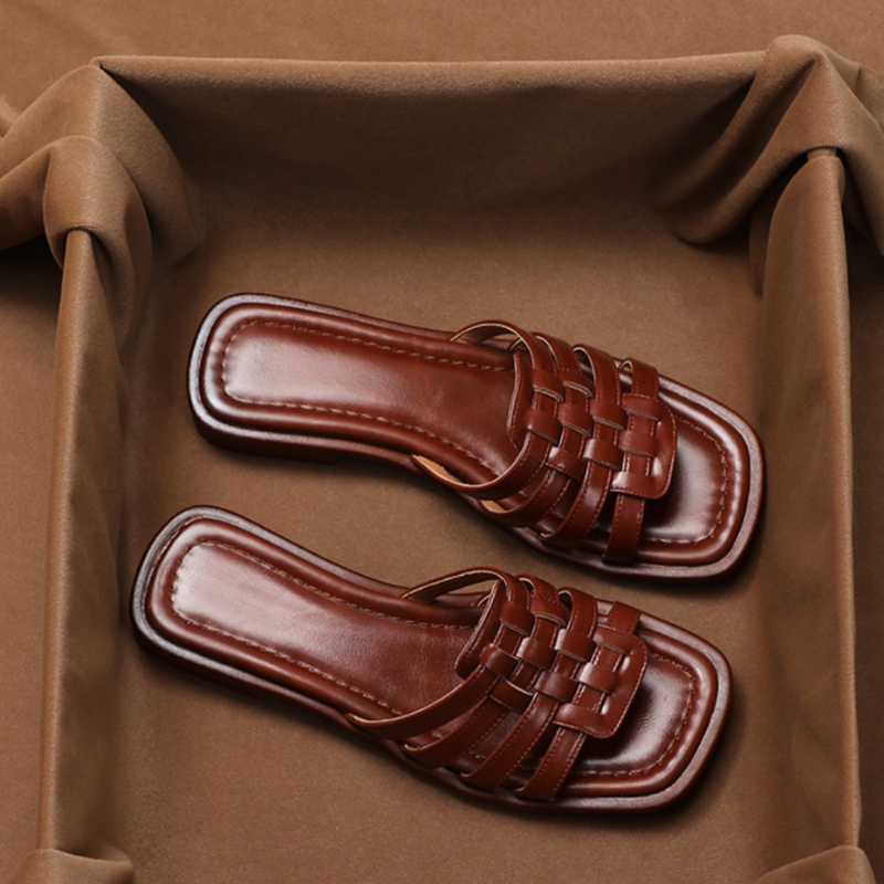 A pair of brown, woven Italian leather slide sandals with a square-toe design, displayed on a soft brown fabric background.