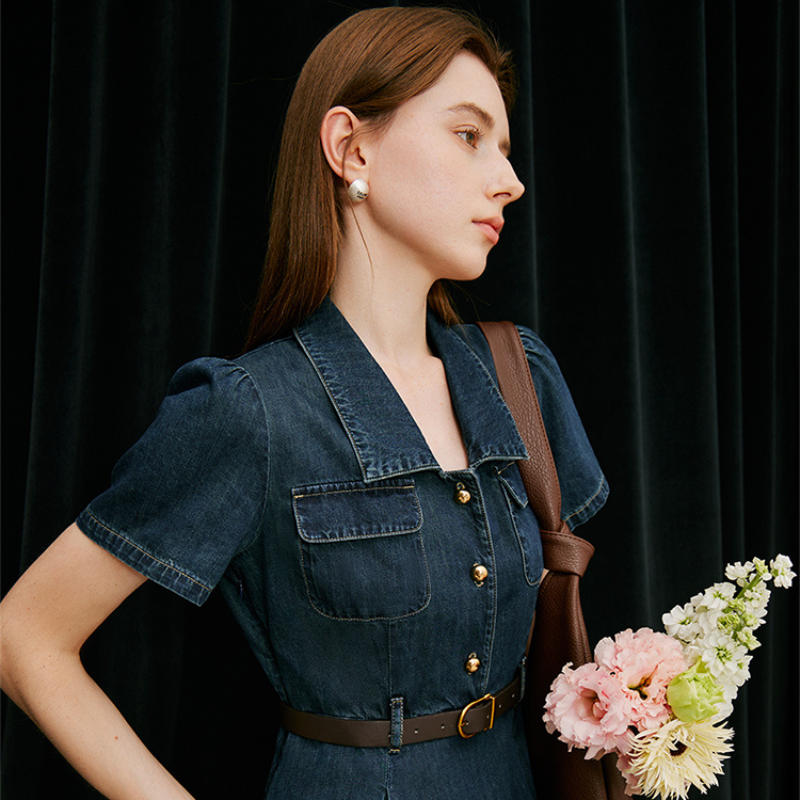 A woman with long brown hair in a denim dress stands against a black curtain, carrying a brown shoulder bag and holding a bouquet of pale pink, yellow, and white blooms—effortlessly embodying chic summer wear.