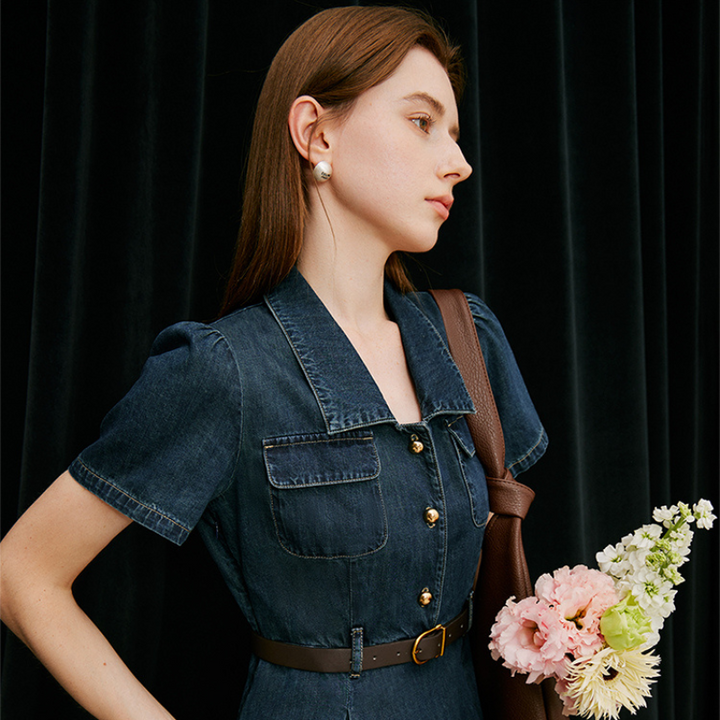A woman with long brown hair in a denim dress stands against a black curtain, carrying a brown shoulder bag and holding a bouquet of pale pink, yellow, and white blooms—effortlessly embodying chic summer wear.