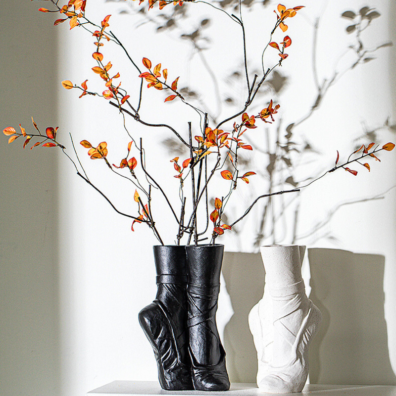 Two ballet-inspired vases shaped like boots—one black, one white—sit on a surface with orange-leaved branches in the black ceramic vase, casting artistic shadows on the wall. Perfect for a touch of contemporary chic décor.