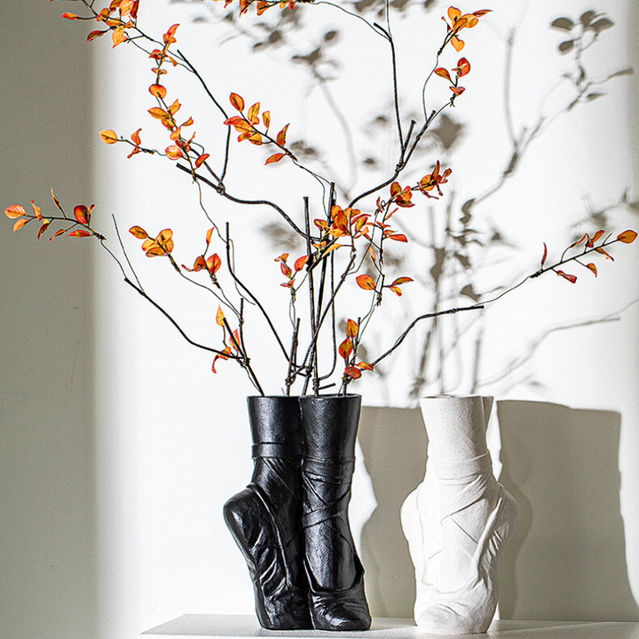 Two ballet-inspired vases shaped like boots—one black, one white—sit on a surface with orange-leaved branches in the black ceramic vase, casting artistic shadows on the wall. Perfect for a touch of contemporary chic décor.