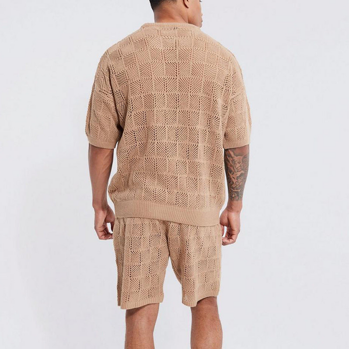 A person is standing facing away from the camera, wearing a matching beige men's 2 piece set with a textured checkered pattern. One arm has a tattoo, and the background is plain white. Perfect for men's summer shorts sets style.