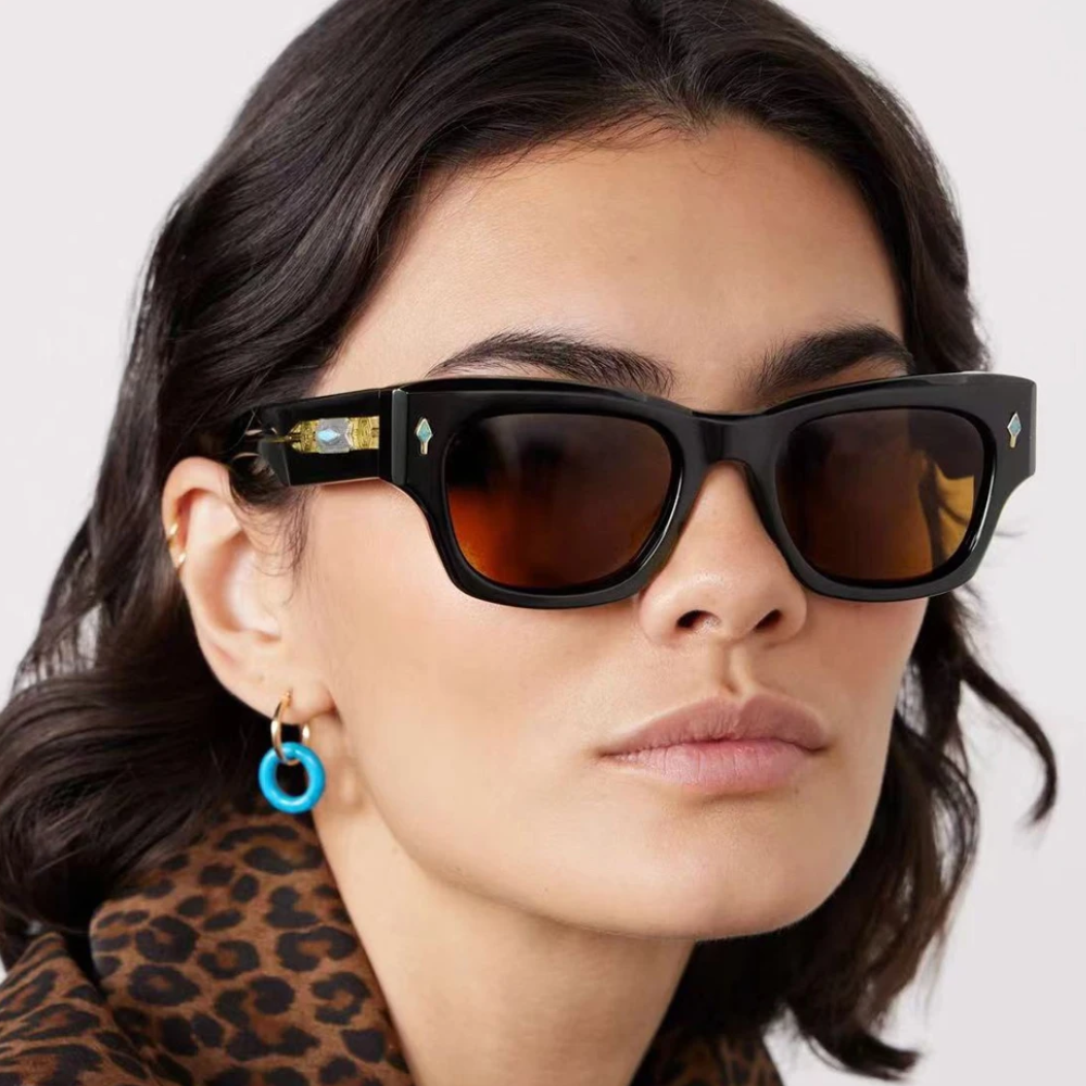 A woman with wavy dark hair wears vintage-inspired sunglasses with brown lenses, a leopard print top, and blue hoop earrings. The background is plain and light-colored.