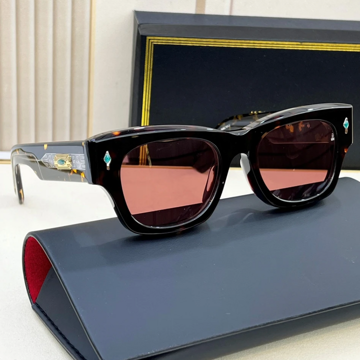 A pair of dark, square-framed vintage sunglasses with red-tinted lenses rests on a blue case. The frame features gold and turquoise decorative details on the sides, and there is a black and gold box in the background.
