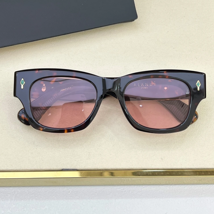 A pair of vintage-inspired tortoiseshell sunglasses with rectangular, pink-tinted lenses is displayed on a cream surface. The frames feature small green and gold decorative accents near the hinges.