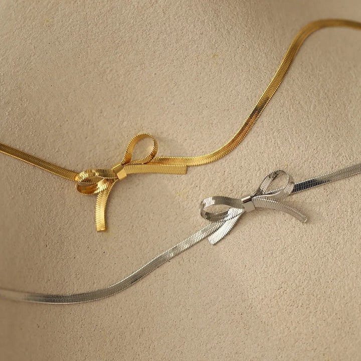A close-up of a beige surface with two metallic bow necklaces, one gold and one silver, each tied into a small bow. This minimalist jewelry piece features neatly crossed ribbons for an elegant touch.