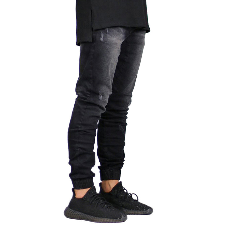 A person wearing black straight tapered leg jeans and black sneakers stands against a white background. The image shows only the legs and lower torso, with the upper body not visible.