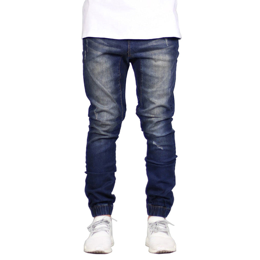 A person wearing dark blue, straight tapered leg, jogger-style jeans with faded details, paired with a white shirt and white sneakers. Only the lower half of the body is visible, giving off a subtle vintage aesthetic.