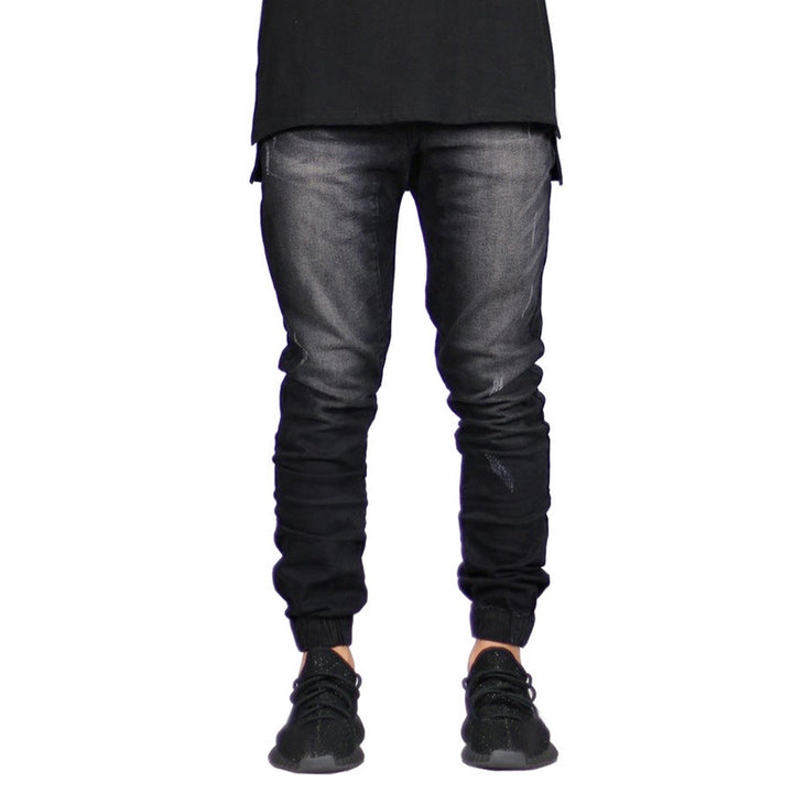 A person wears black jogger jeans with a faded effect and straight tapered leg, paired with a long black shirt and black sneakers. The image is cropped above the shoulders, highlighting a modern look with vintage aesthetic.