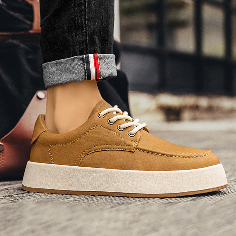 A person wearing cuffed dark jeans and tan men’s leather casual shoes with thick white soles stands on a textured surface, with a blurred background.