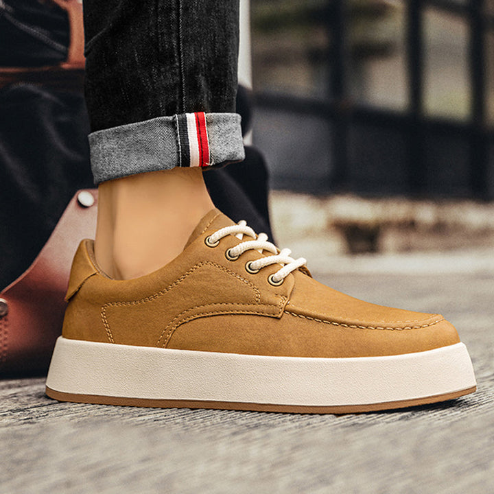 A person wearing cuffed dark jeans and tan men’s leather casual shoes with thick white soles stands on a textured surface, with a blurred background.