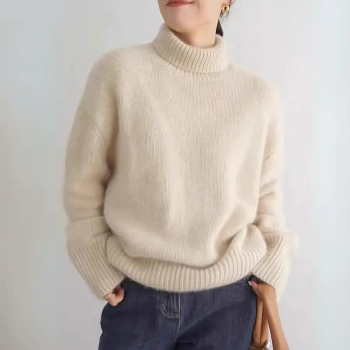 A person wearing a cream-colored, oversized fit chunky knit turtleneck sweater with blue jeans, standing against a plain light background. Their hands are in their pockets and their face is partially out of frame.