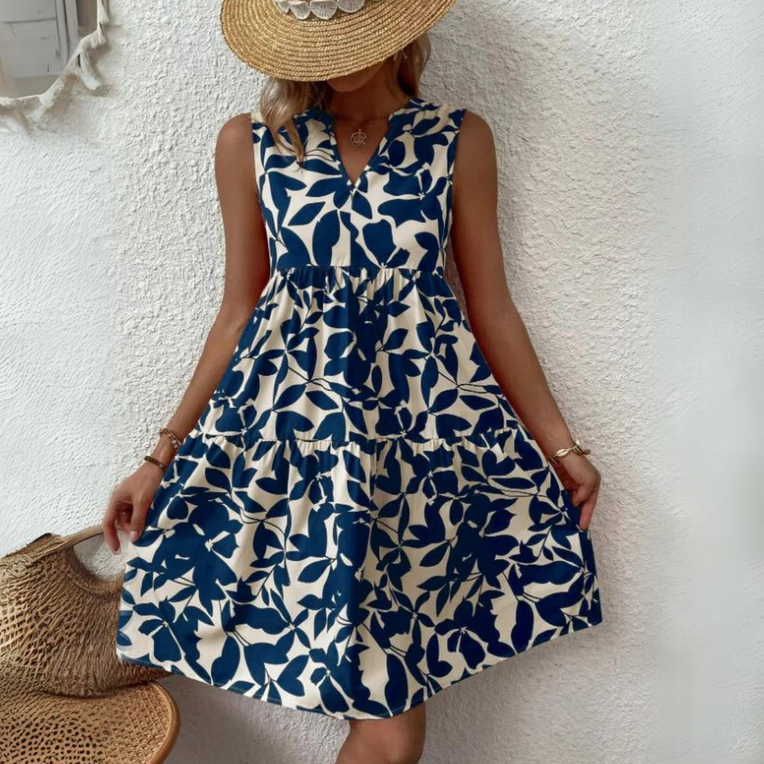 A woman in a sleeveless dress made of lightweight breathable fabric with a blue and white leaf pattern stands against a textured white wall. She wears a wide-brimmed straw hat, obscuring her face, and holds the sides of her dress—perfect for a summer wardrobe.