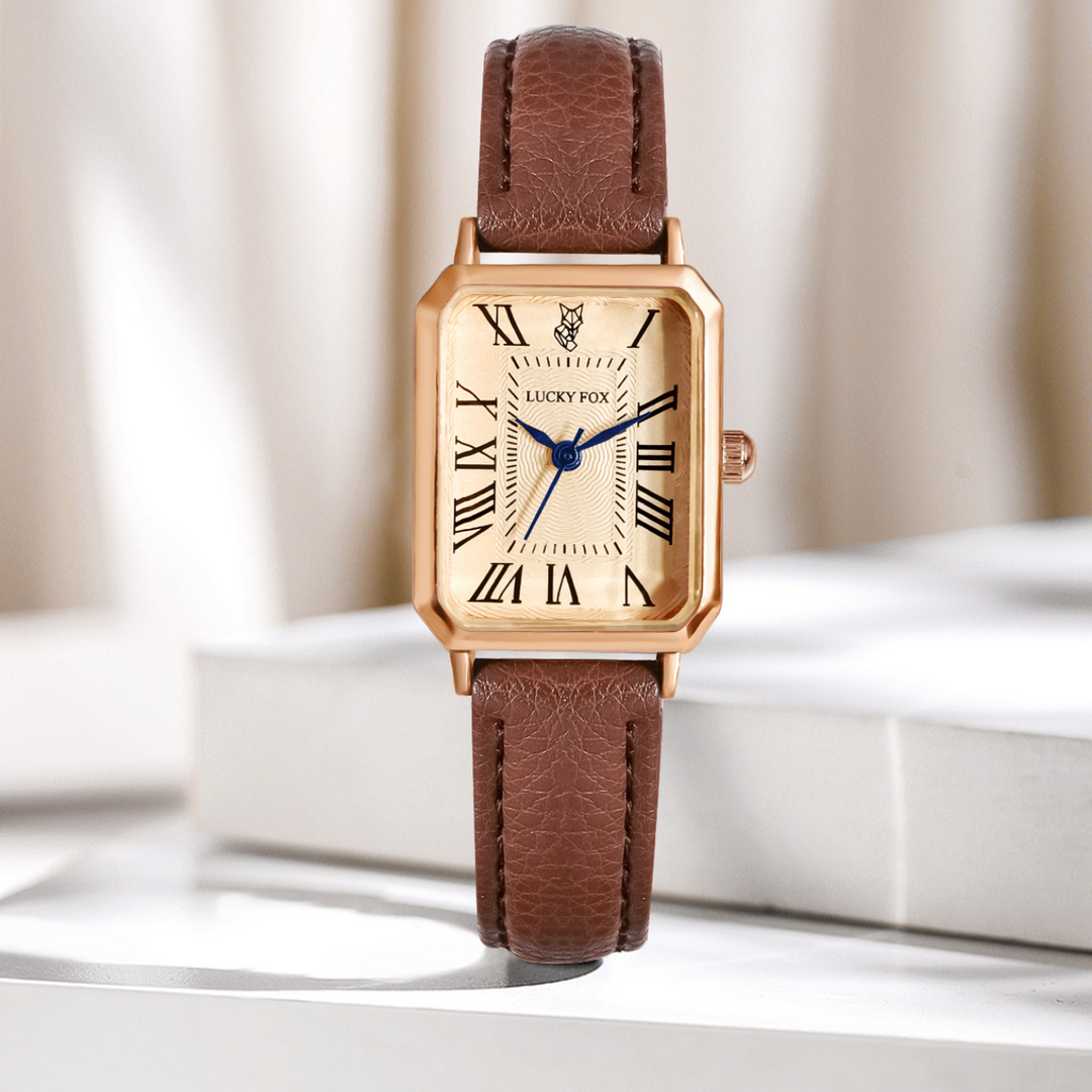 A rectangular face watch with a genuine leather strap, Roman numerals, and blue clock hands stands upright on a white surface against a softly blurred beige background.