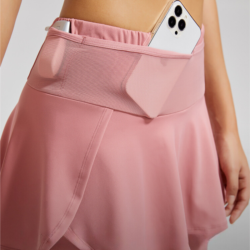 A person wearing a pink athletic skirt with stretch storage pockets and a mesh waistband pocket, holding a smartphone on the right side of their waist.
