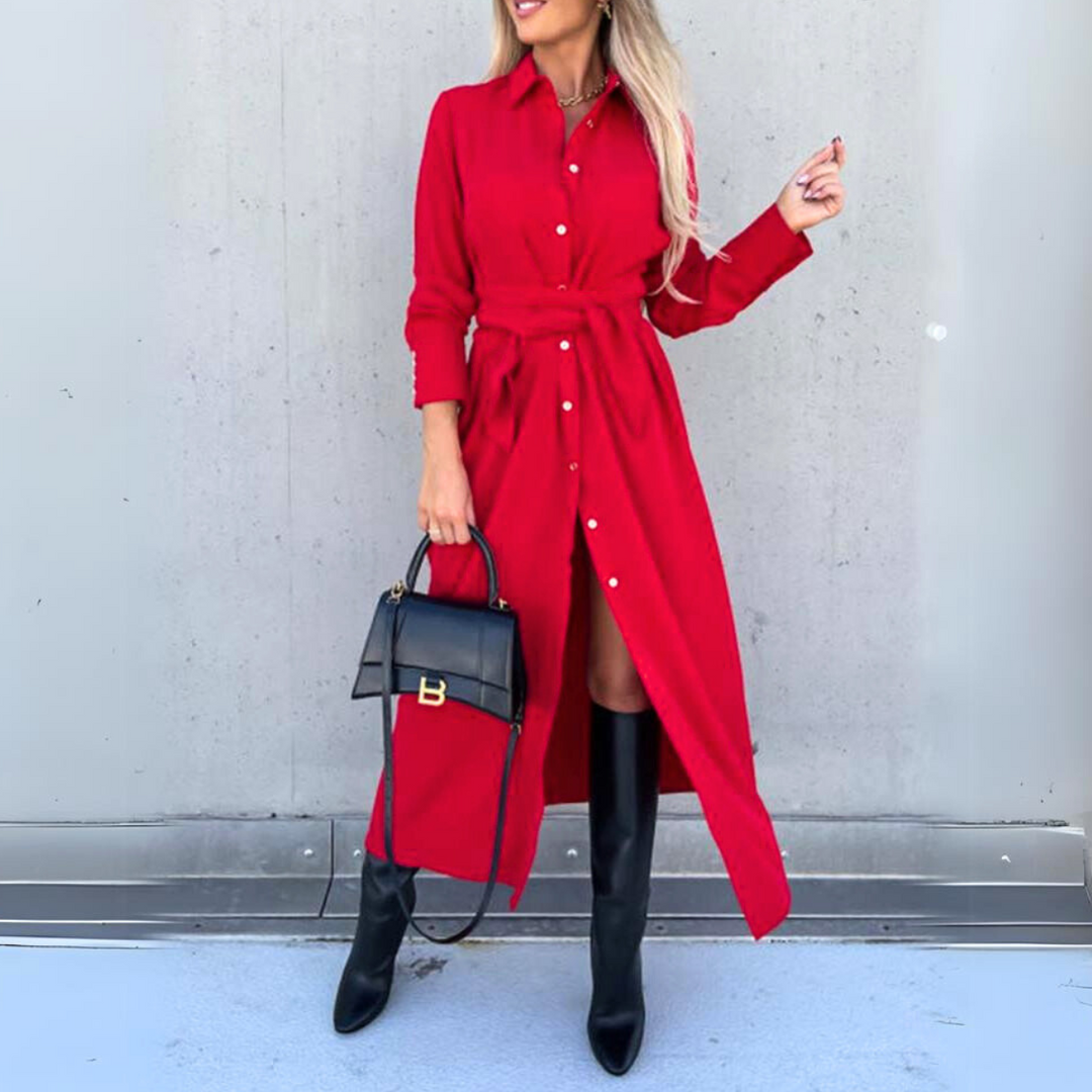 A woman in a high fashion, long-sleeve wrap dress and knee-high black boots holds a black handbag, standing against a plain light gray wall. Her long blonde hair is styled loosely.