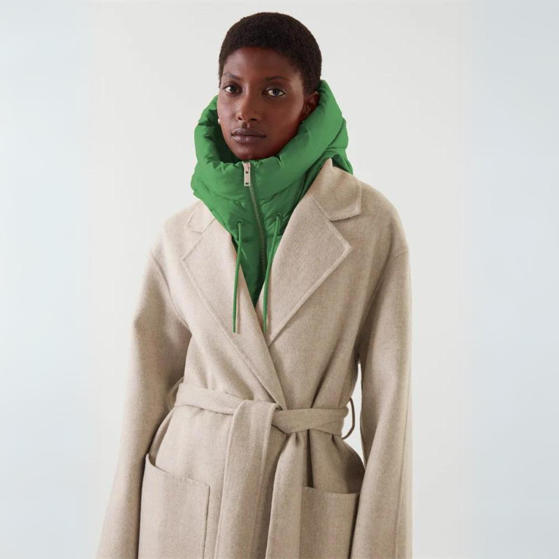 A person wearing a beige belted coat crafted from durable material over a green puffer vest with a high collar, standing against a plain light background.