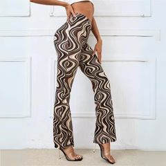 A person poses sideways, wearing trendy statement pants with high-waisted, brown and beige swirl patterns and clear high-heeled shoes. The cropped top reveals their midriff. The background features a white wall with decorative panels.