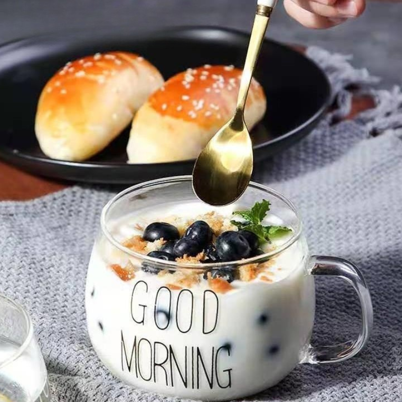 A glass mug with a clear design labeled GOOD MORNING, filled with yogurt, blueberries, and granola, sits on a table. A hand holding a gold spoon hovers above it. In the background, two bread rolls rest on a black plate.