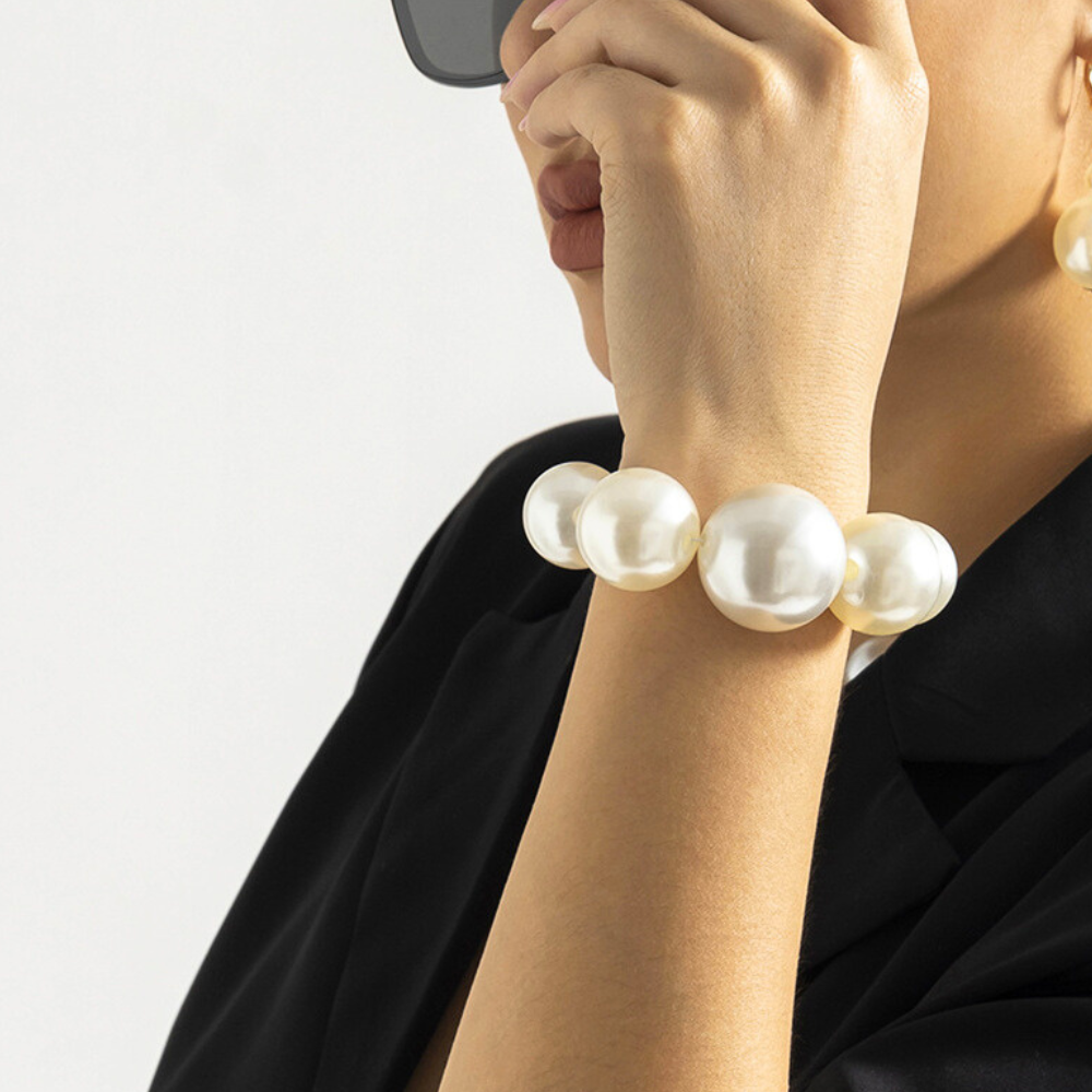 A person wearing black sunglasses and a black outfit poses with their hand near their face, showing off a chunky pearl jewelry set and matching earrings—bold statement jewelry that elevates classic streetwear accessories.