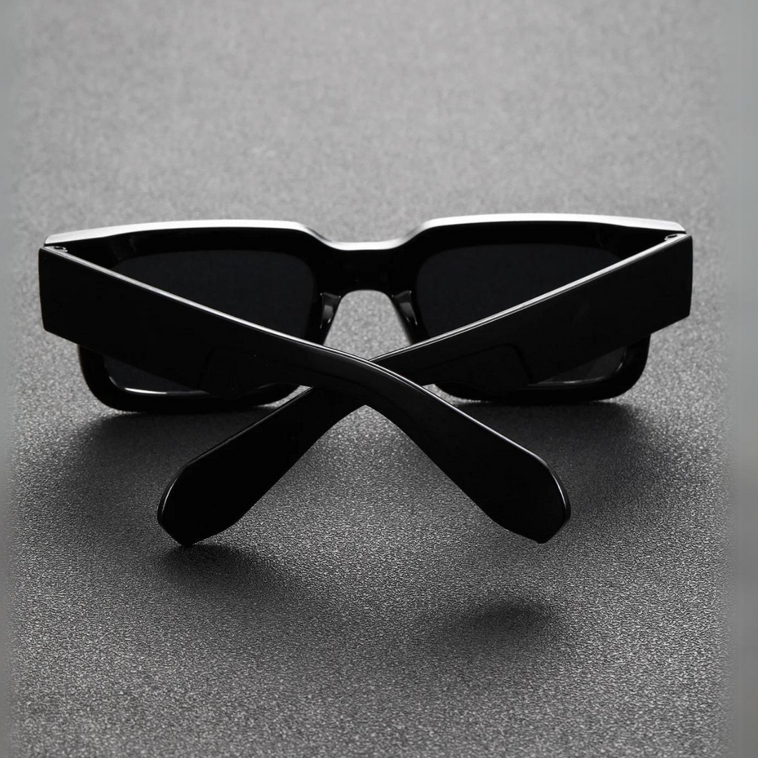A pair of black lightweight sunglasses with thick frames is placed upside down on a dark textured surface, with the arms crossed in an X shape in front of the lenses.