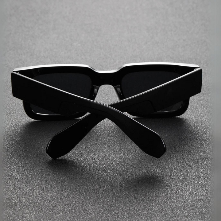 A pair of black lightweight sunglasses with thick frames is placed upside down on a dark textured surface, with the arms crossed in an X shape in front of the lenses.