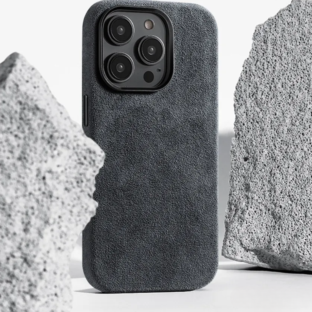 A smartphone with a suede finish, protective design case stands upright between two large, rough, light gray stones on a white background.