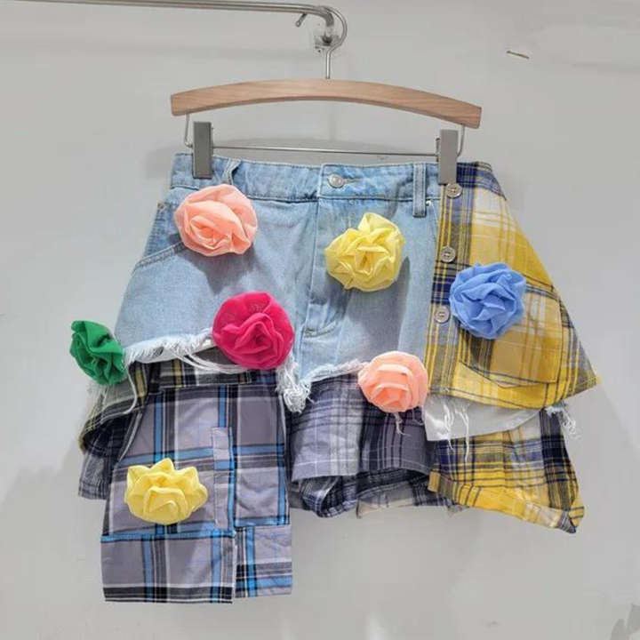 A patchwork skirt made from denim and plaid, decorated with large, colorful fabric flowers in yellow, pink, peach, and blue, hangs on a wooden hanger against a plain white background—perfect for an eclectic fashion statement.