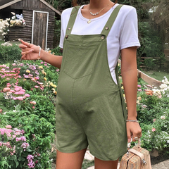 A person wearing comfortable maternity overalls and a white t-shirt stands in a garden filled with colorful flowers, holding a woven handbag. A wooden shed is visible in the background.